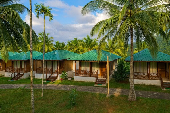 A row of bungalow-style accommodations with green roofs, surrounded by palm trees and lush greenery. - Rainforest Resort (Port Blair)