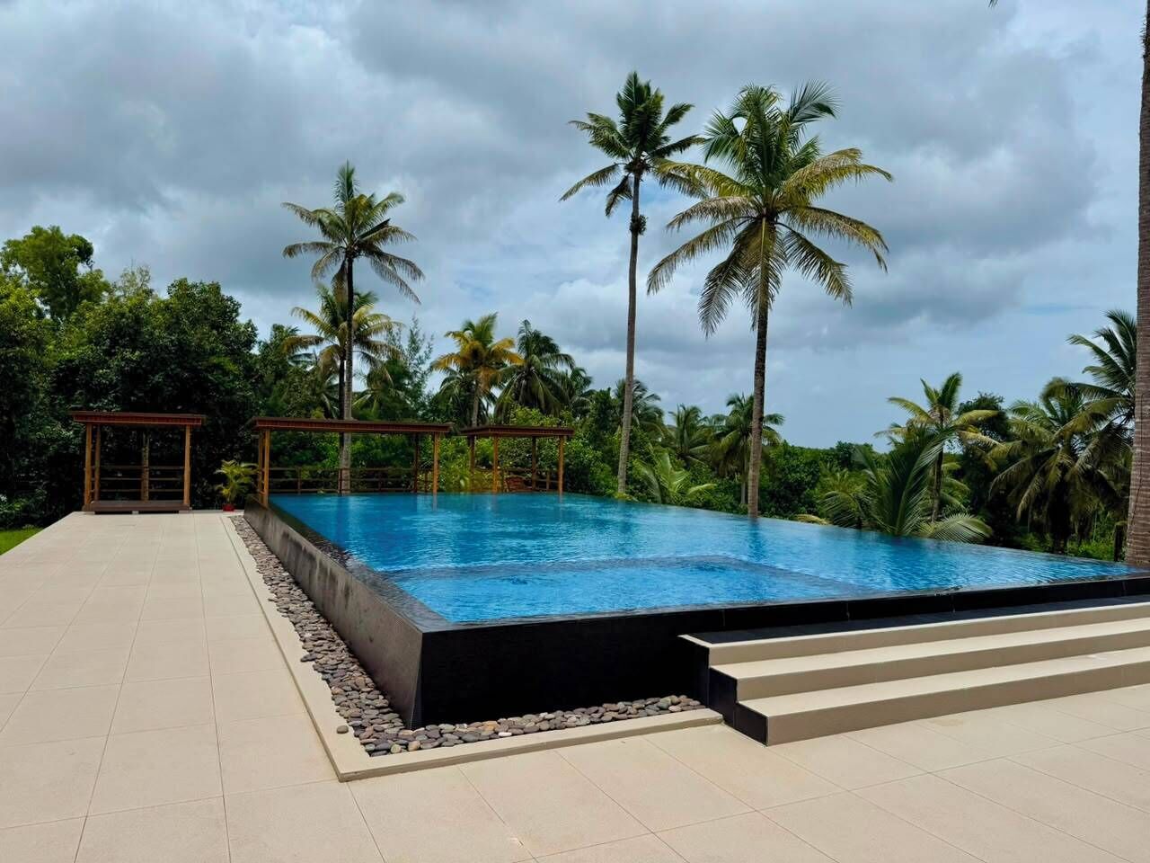 Una piscina infinita con superficie de agua azul cristalina, rodeada de palmeras y una zona de estar cubierta.