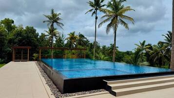 Una piscina infinita con superficie de agua azul cristalina, rodeada de palmeras y una zona de estar cubierta.