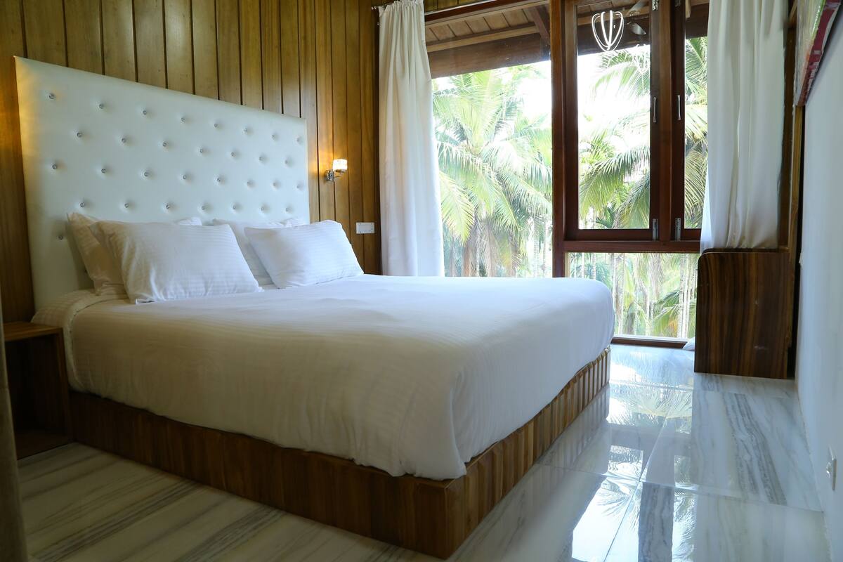 Uma cama grande com cabeceira de madeira e roupa de cama branca, uma mesa de cabeceira e vista para palmeiras pela janela.