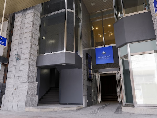 A modern hotel entrance with a glass facade, a blue sign indicating the name, and a staircase leading to the lobby.