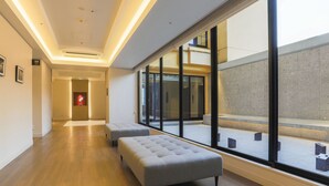 A modern hotel corridor with wooden flooring, grey upholstered seating, and large windows offering views of the exterior.