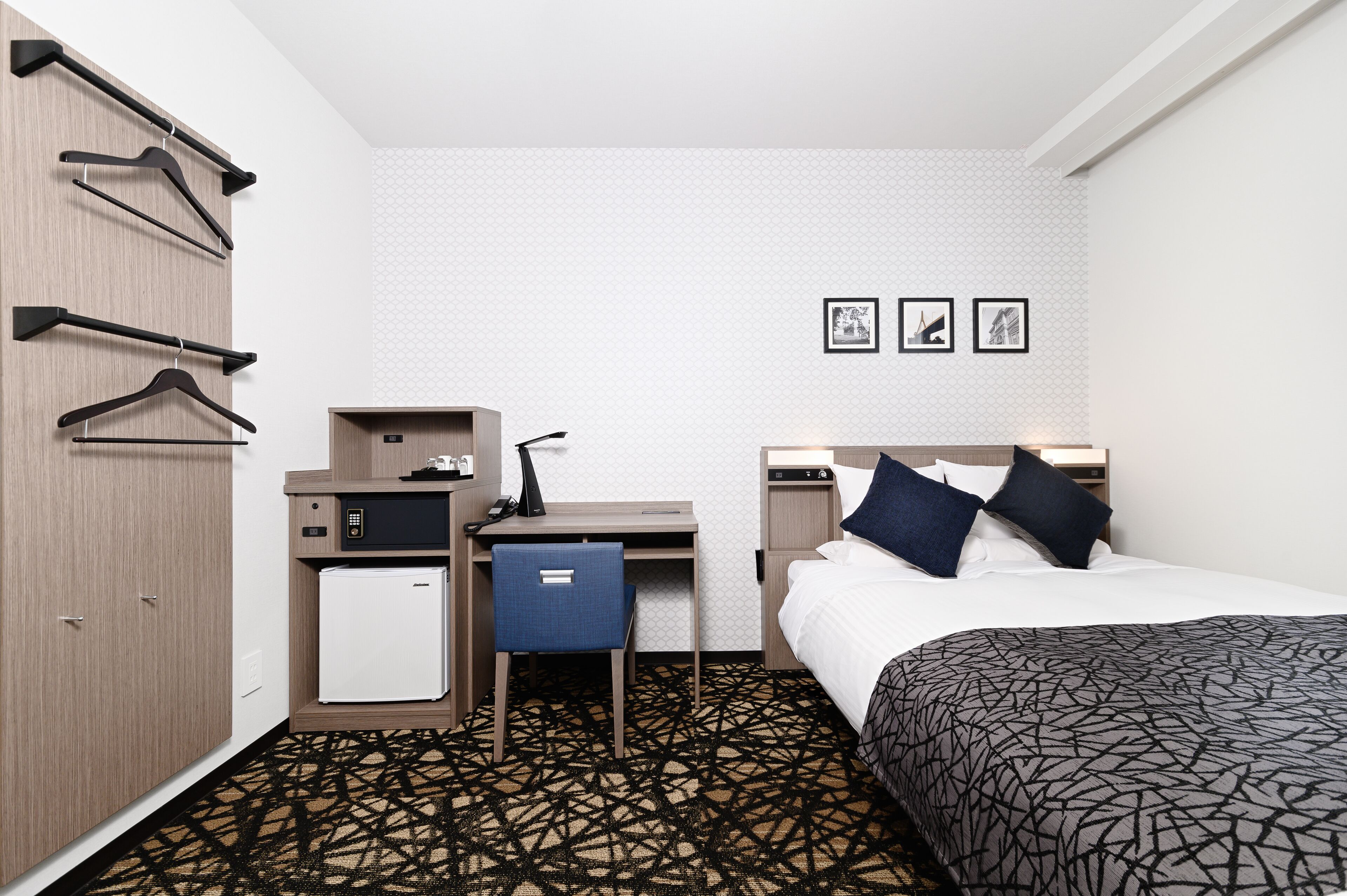 A hotel room with a bed, a desk with a chair, a wardrobe, and a small refrigerator. There are three framed pictures on the wall.