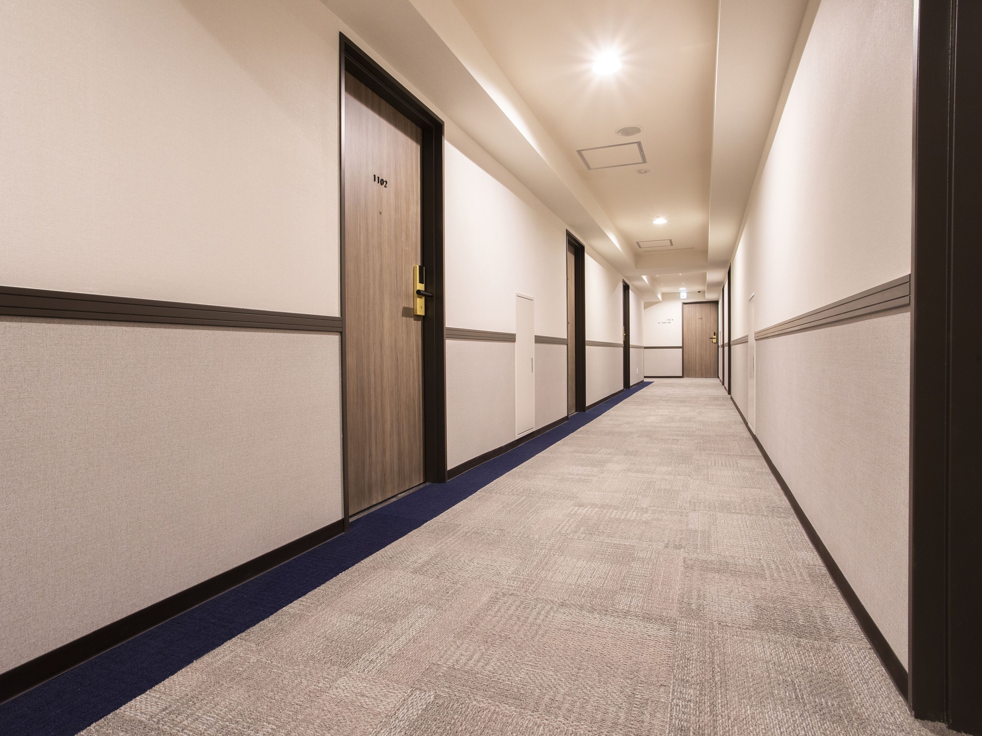 Un long couloir étroit, typique d’un hôtel, avec des revêtements de sol en moquette et des portes de part et d’autre.