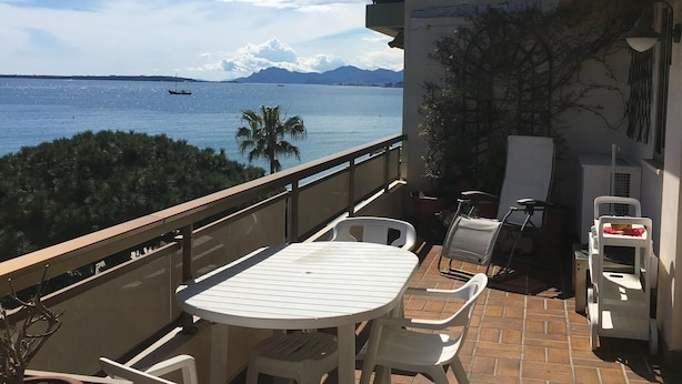 A balcony with a table and chairs, overlooking a body of water with a boat and mountains in the distance. - JUAN LES PINS STUDIO SEA VIEW (Juan Les Pins)