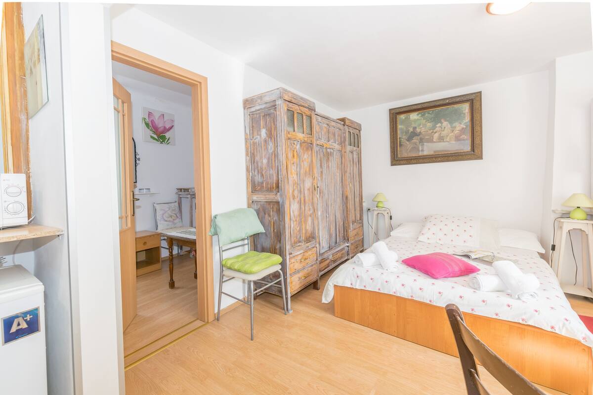 A bedroom with a wooden wardrobe, a bed with floral bedding, a chair, and a desk with a lamp.