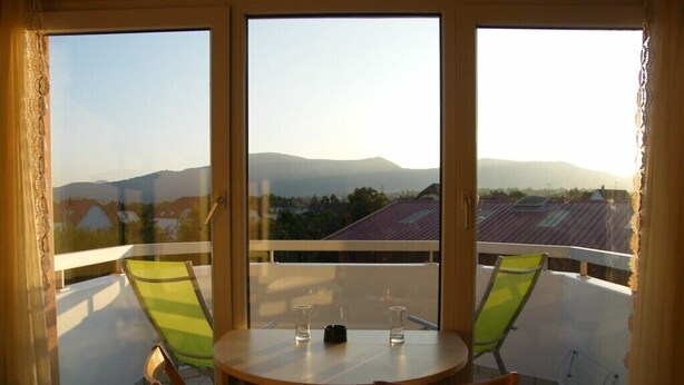 Un balcon avec une table ronde en bois et deux chaises vertes, donnant sur un quartier résidentiel avec des maisons et des montagnes.