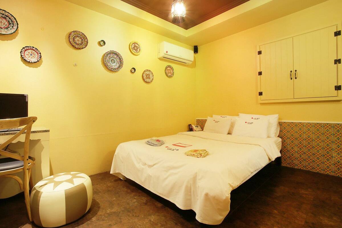 Een kamer met een bed, een bureau en een stoel. Er hangen decoratieve borden aan de muur en er is een airconditioner.
