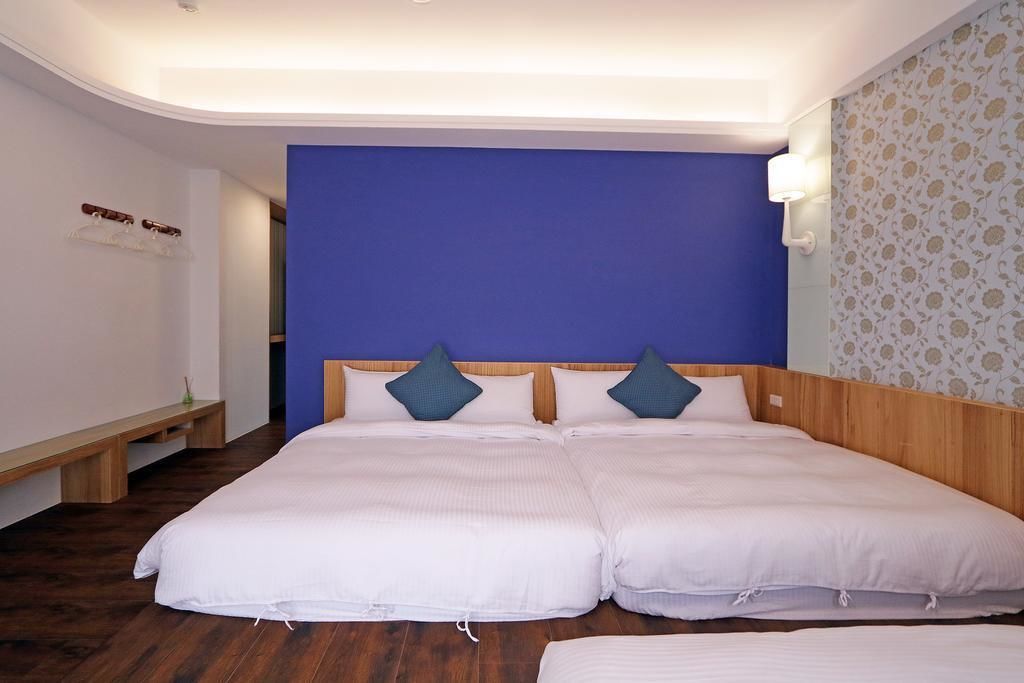 Un lit double avec une literie blanche et deux oreillers bleus, un banc en bois et un mur orné d’un motif floral.