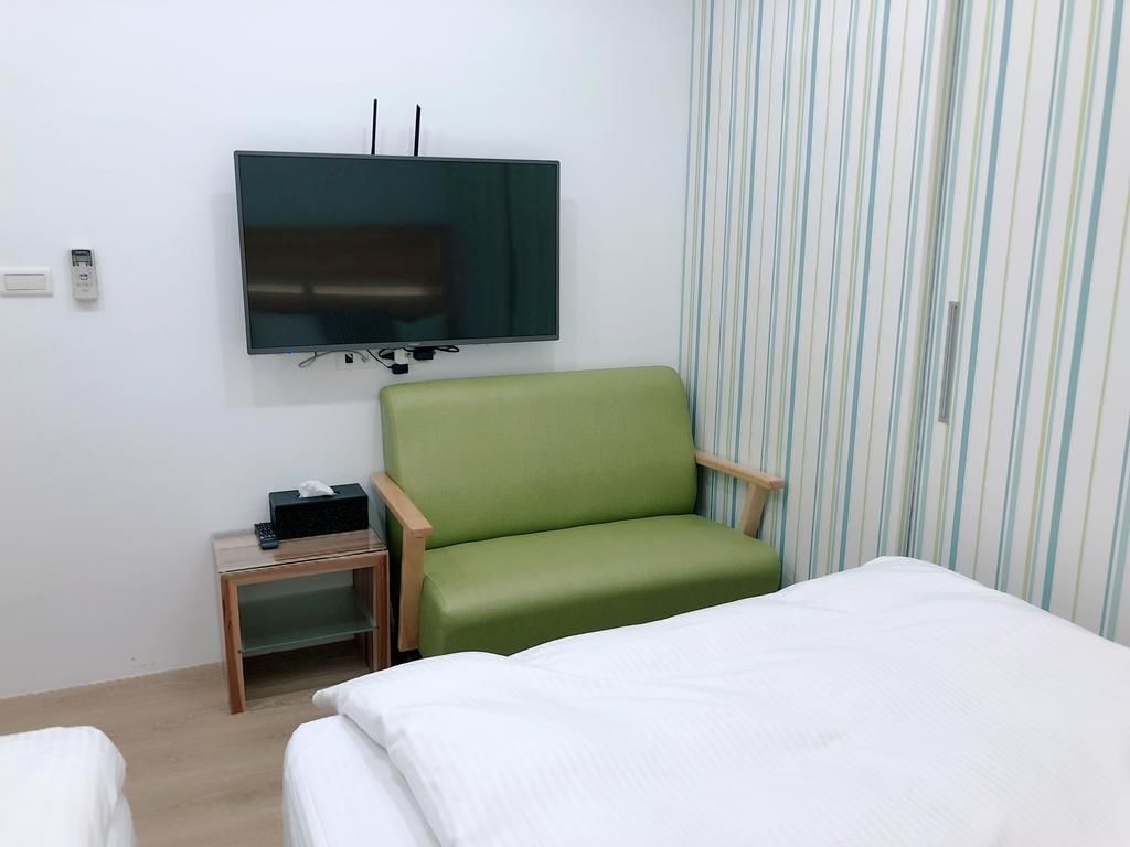 Una habitación de hotel con una cama, un sillón verde, un televisor montado en la pared y una cortina a rayas.