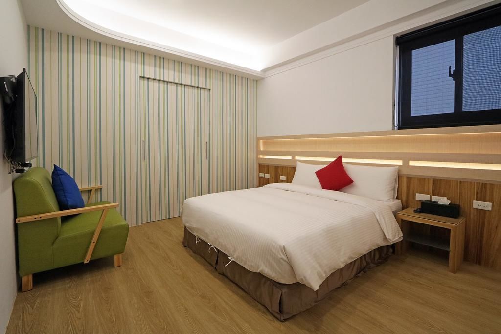 Een hotelkamer met een bed, een groene fauteuil, een aan de muur bevestigde televisie en een raam met zonwering.