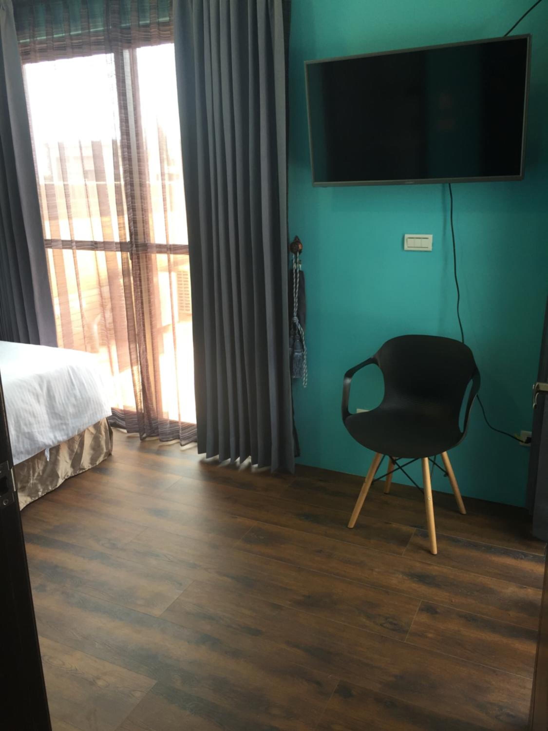 A room with a bed, a chair, a TV mounted on the wall, and a window with curtains.