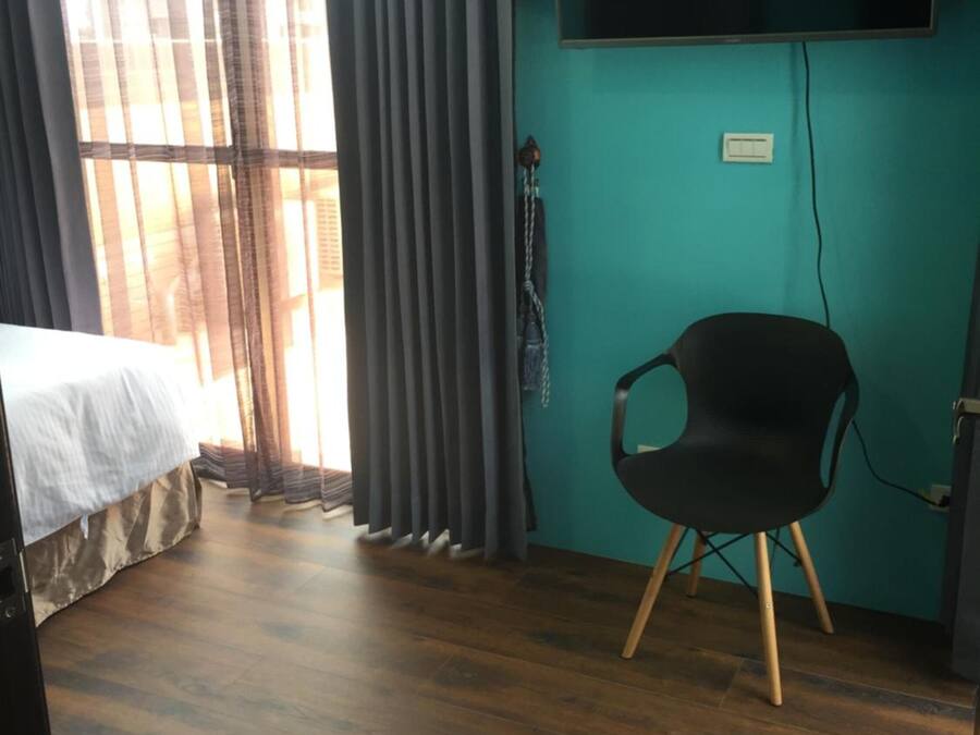 A room with a bed, a chair, a TV mounted on the wall, and a window with curtains.