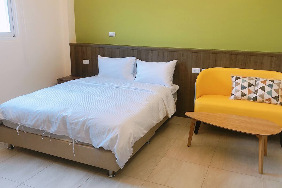 A bedroom with a bed, a nightstand, a wooden headboard, a yellow sofa, and a patterned pillow.