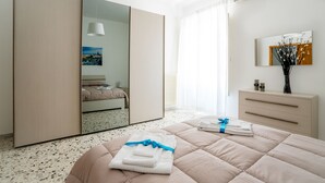 Une chambre moderne avec un grand miroir, un lit sur lequel sont soigneusement pliées des serviettes, une commode et une fenêtre ornée de rideaux légers.