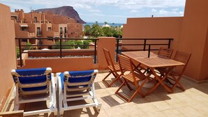 A balcony with two blue lounge chairs and a wooden table, overlooking a mountainous landscape and the sea. - La Tejita - with two terraces depending on the direction of wind and sun. (Los Abrigos)