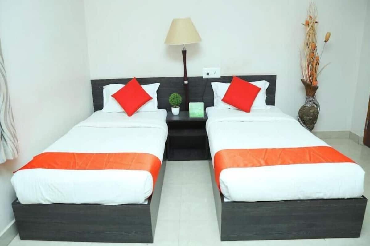 Habitación de hotel con dos camas, cada una con ropa blanca y detalles naranja, una mesita de noche con una lámpara, una planta pequeña y un florero con flores secas.