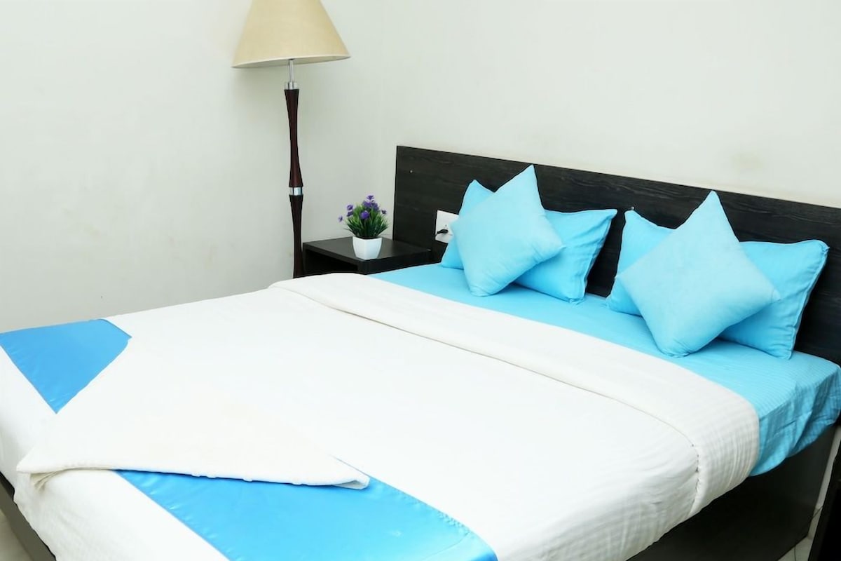 Una cama con una colcha blanca y azul, un cabecero oscuro y una mesita de noche pequeña con una lámpara y una planta en maceta.