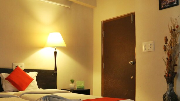 Quarto de hotel com cama, mesa de cabeceira, luminária, porta e um vaso com flores secas.