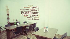 Interior de restaurante com mesa e cadeiras de madeira, uma nuvem de palavras na parede e um vaso com plantas secas.