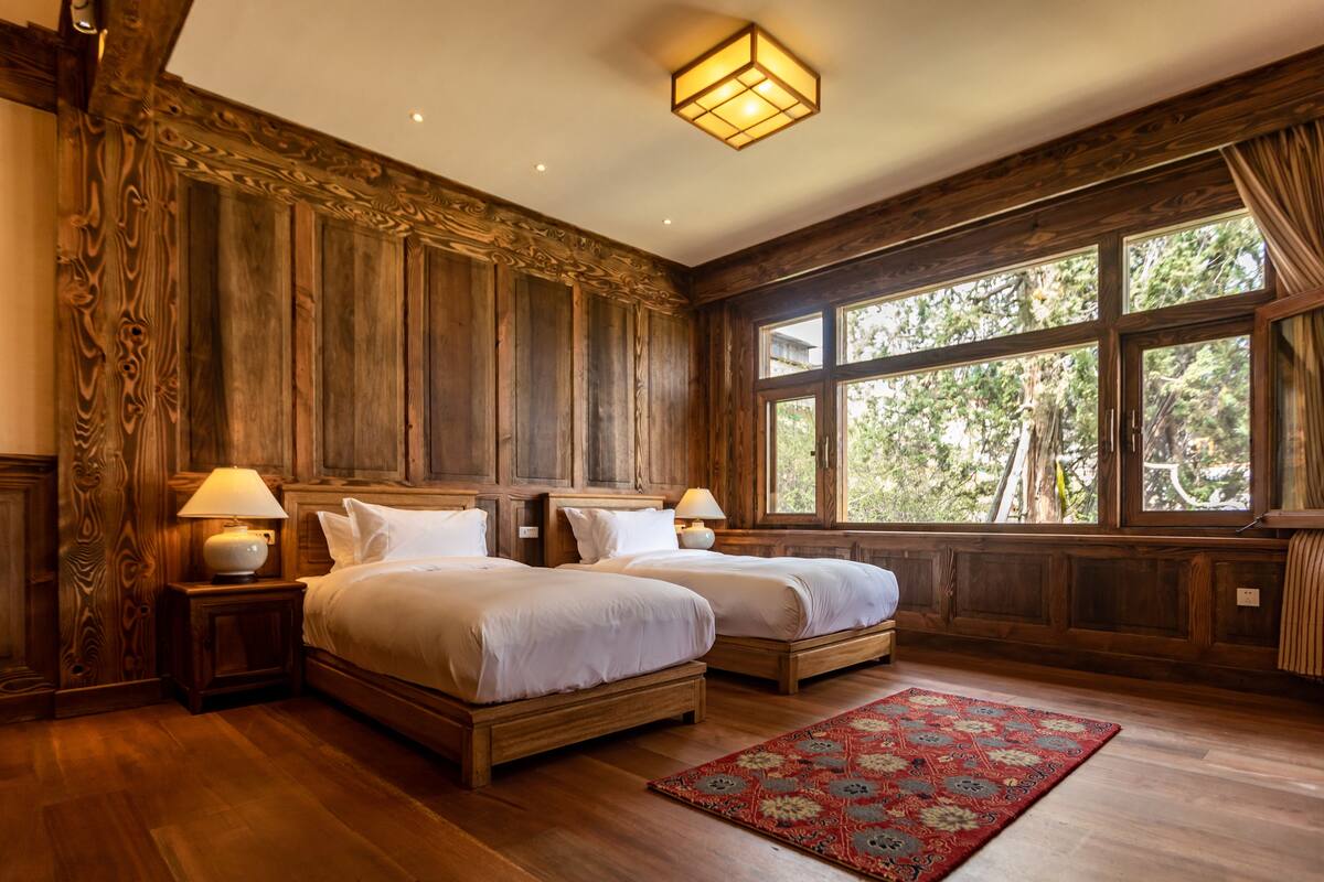 Una habitación con dos camas de madera, un tapete rojo estampado y ventanas amplias con vistas a árboles.