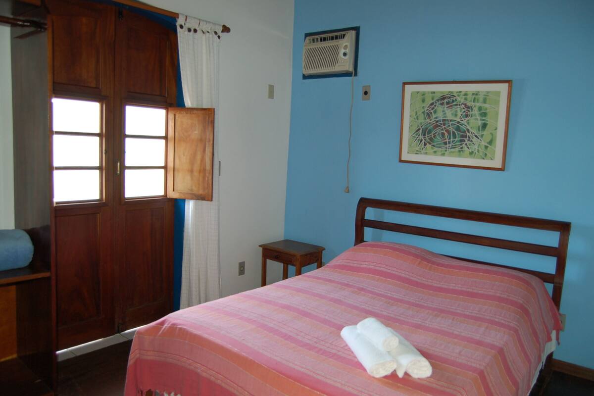 A bedroom with a wooden bed, a small bedside table, a wall-mounted heater, and a window with curtains.