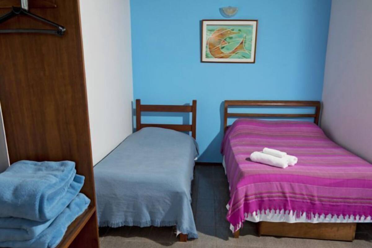 A bedroom with two single beds, one with a blue blanket and the other with a pink blanket. There is a wooden wardrobe on the left and a framed picture on the wall.