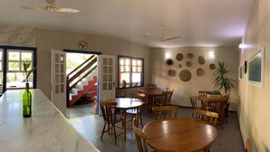 A dining area with wooden tables and chairs, a bar area with a green bottle, and a staircase leading to a red room. - Pousada Manacá (Paraty)