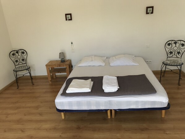 Un letto rifatto con cuscini e asciugamani piegati, un piccolo comodino in legno con una lampada e due sedie in metallo.
