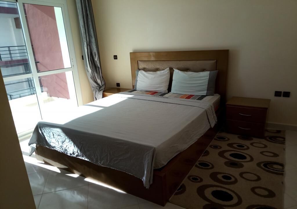 Een hotelkamer met een bed, nachtkastje, lade, een kamerbreed tapijt met dessin en een raam met gordijnen.