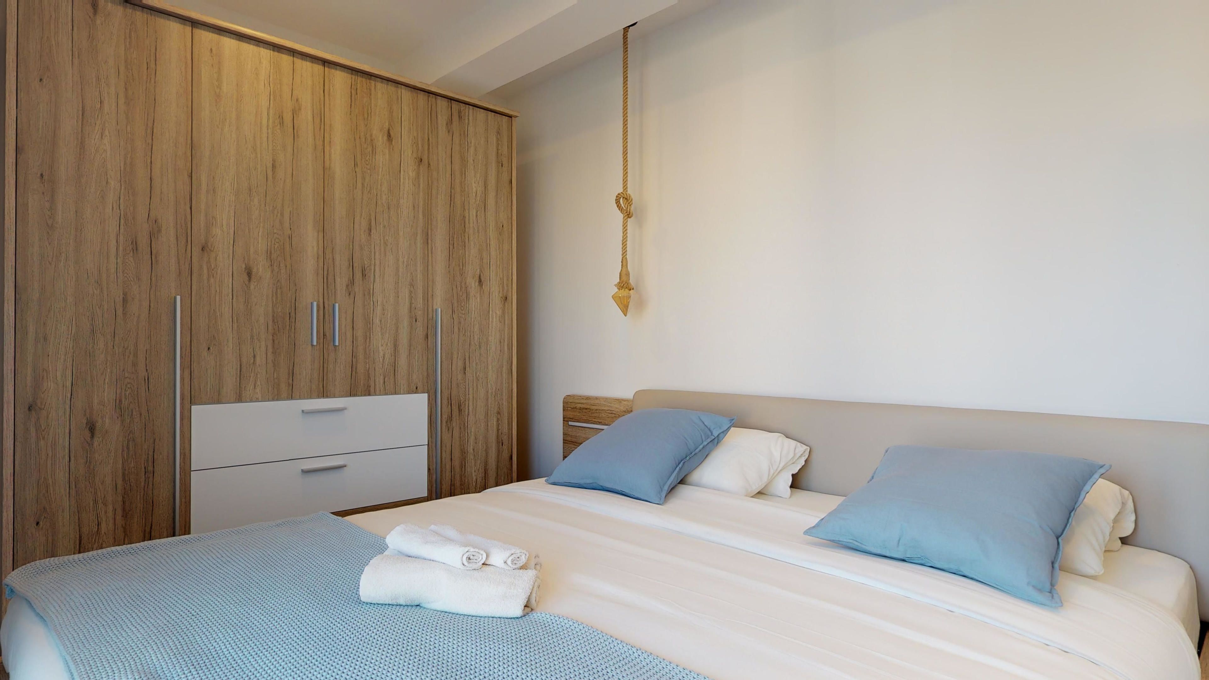 A bedroom with a bed, two pillows, a bedside table, a wardrobe, and a hanging light fixture.