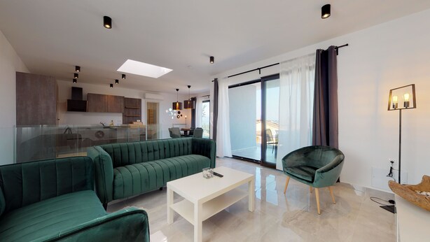 A modern living room with a green sofa, a white coffee table, and a green armchair. There is a kitchen area in the background with a dining table and chairs. - Istrian Pearl (Rasa)