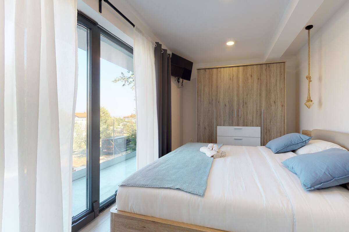Une chambre avec une grande fenêtre, un lit avec des oreillers bleus, une armoire en bois et un téléviseur à écran plat fixé au mur.