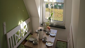 Un coin repas avec une table dressée pour quatre personnes, un banc blanc, une fenêtre donnant sur un champ et un luminaire suspendu.