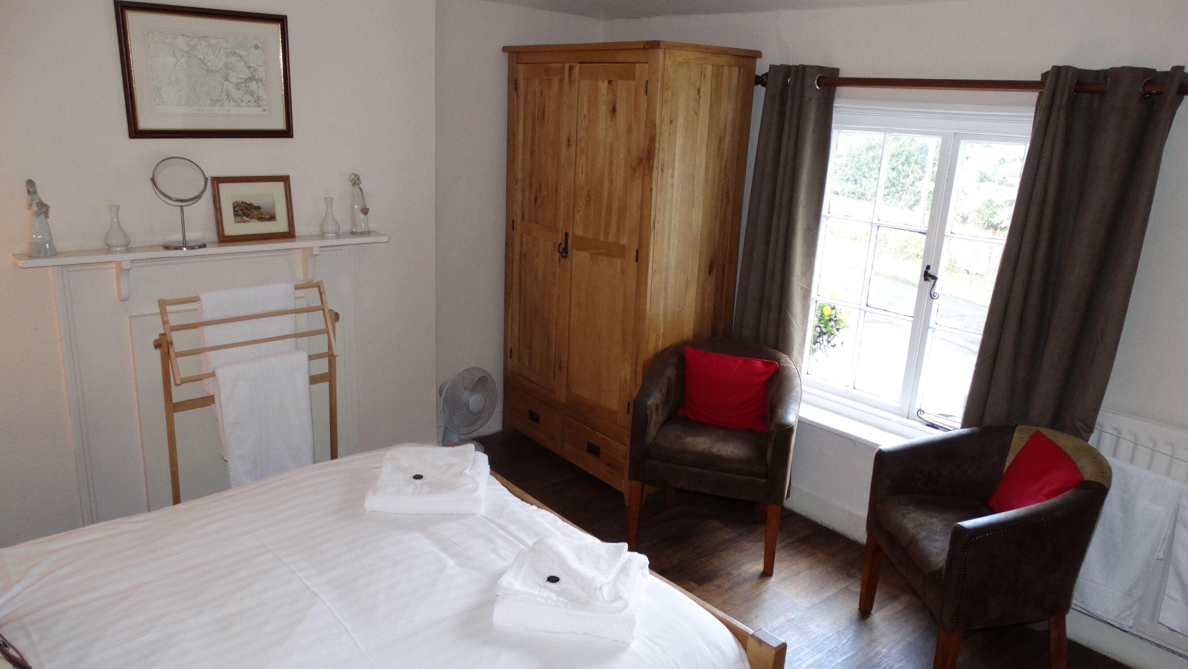 Een slaapkamer met een bed, een houten garderobe, een stoel met een rood kussen, een klein tafeltje met een spiegel en een raam met gordijnen.