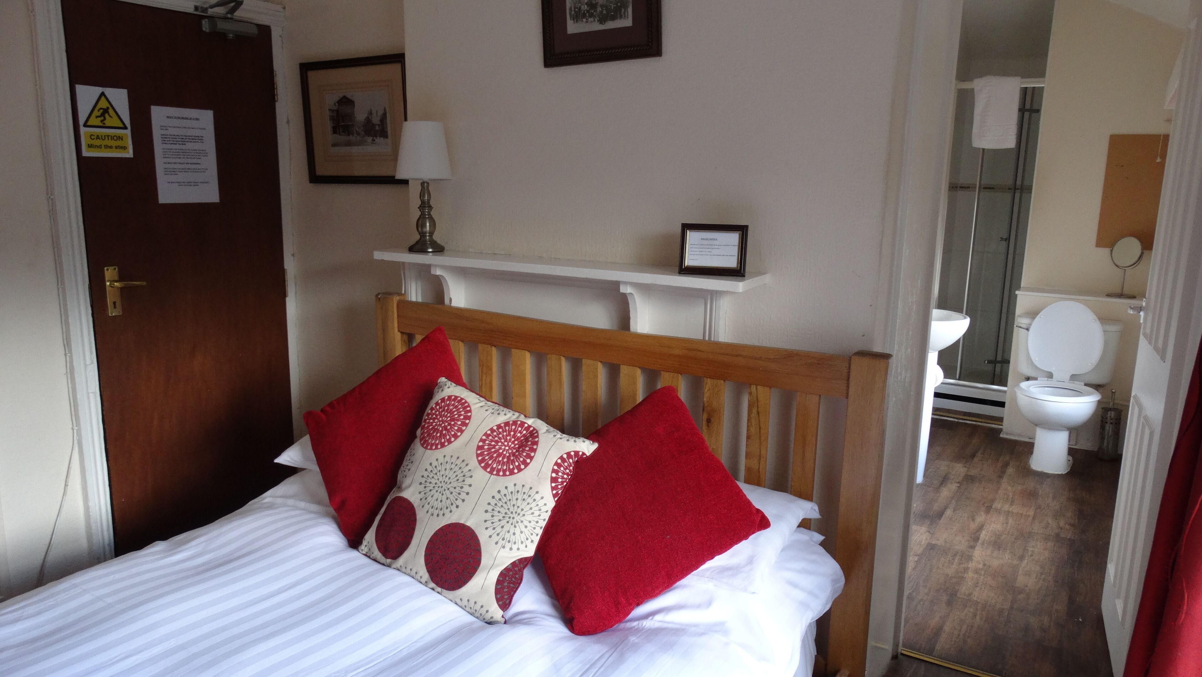 A bedroom with a wooden headboard, a bed with white and red bedding, a bedside table with a lamp, and a framed picture on the wall.