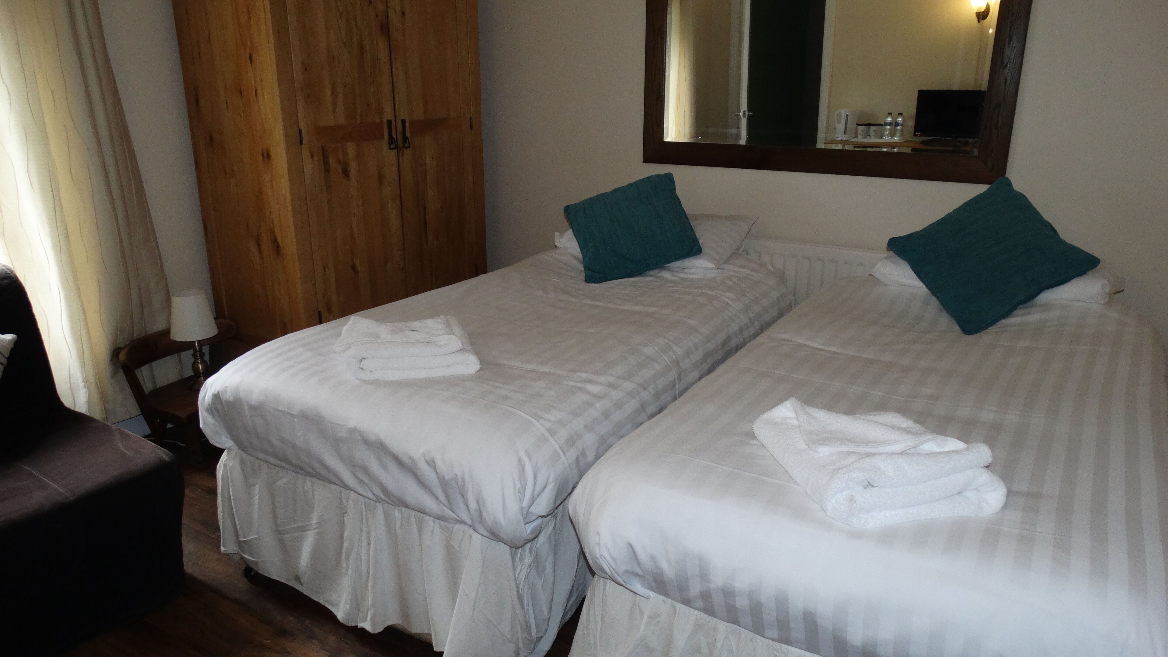 A hotel room with two beds, each with a folded towel, a wooden wardrobe, a mirror, and a table with a lamp.