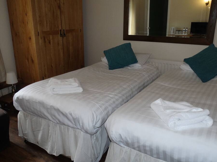 A hotel room with two beds, each with a folded towel, a wooden wardrobe, a mirror, and a table with a lamp.