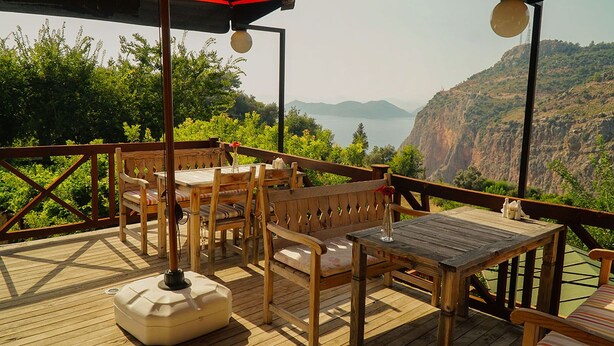 A wooden deck with outdoor furniture, a table, and a bench, overlooking a mountainous landscape and a body of water.