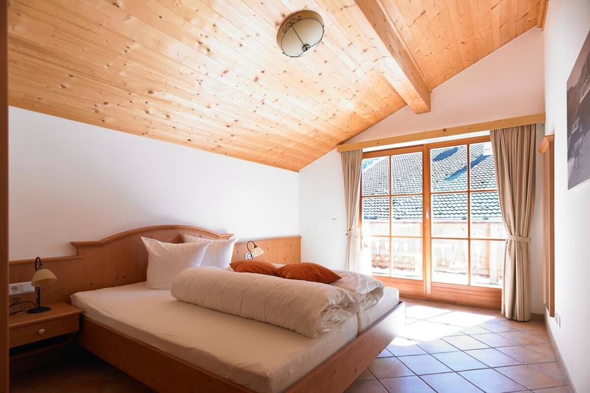 Une chambre avec un plafond en bois, une grande fenêtre donnant sur un beau paysage, un lit avec des oreillers et une table de chevet avec une lampe.