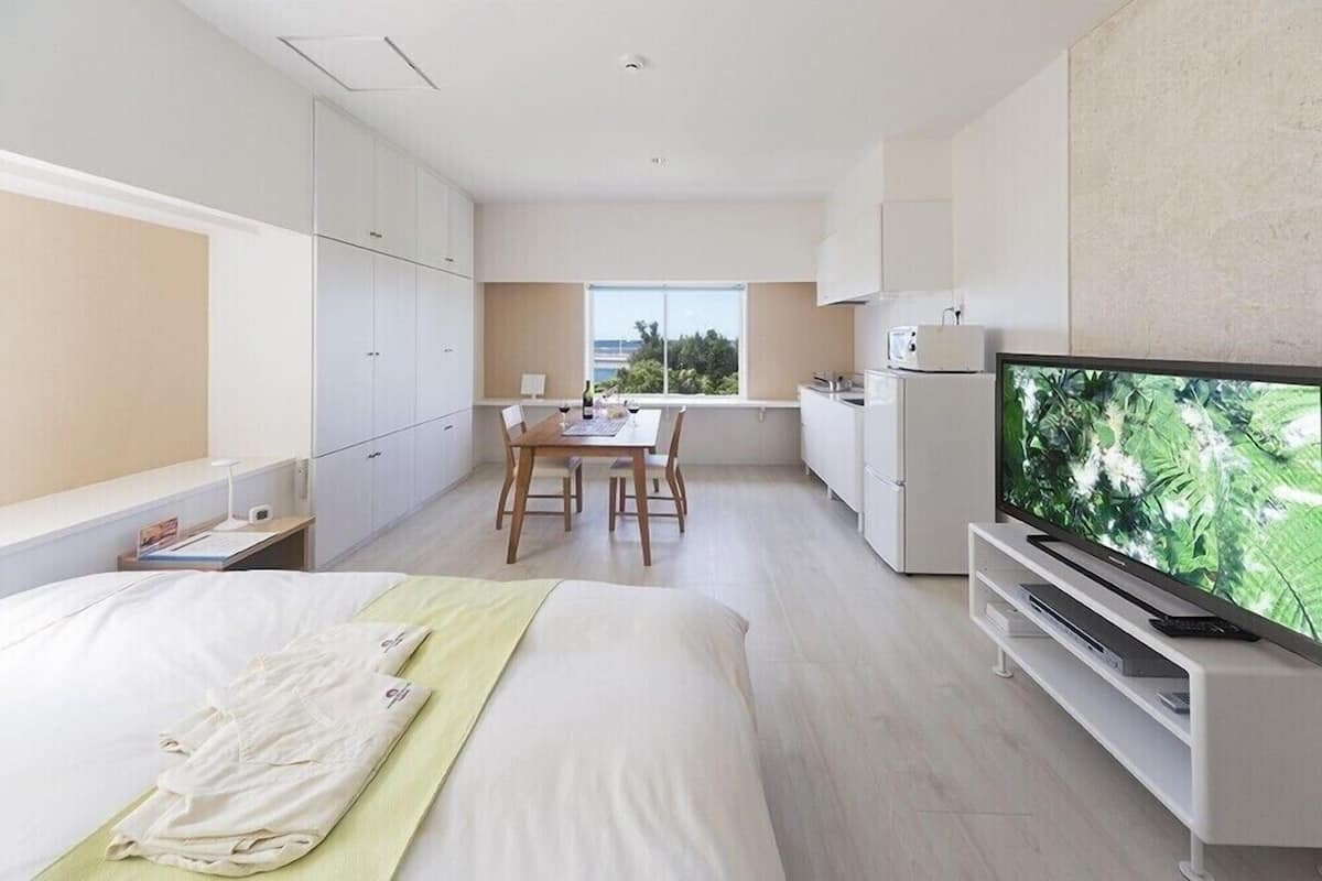 A modern hotel room with a bed, a dining table with chairs, a kitchen area, and a television.