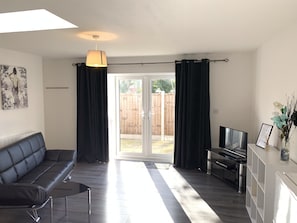 A modern living room with a black leather sofa, a glass coffee table, a flat-screen TV, and a dining area with a white cabinet. - Grand Central Budget Modern Apartments (Derby)