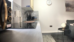 Eine moderne Küche mit einem Edelstahl-Kühlschrank, einer Kaffeemaschine und einem Wasserkocher. Ein Waschbecken mit schwarzem Wasserhahn und ein schwarzes Metallregal sind vorhanden. An der Wand hängt eine Uhr, und ein kleiner runder Tisch mit einer Lampe ist zu sehen.