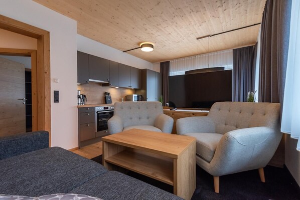 Una sala de estar moderna con techo de madera, sillas tapizadas en gris, una mesa de centro de madera y una zona de cocina con armarios grises y un horno empotrado.