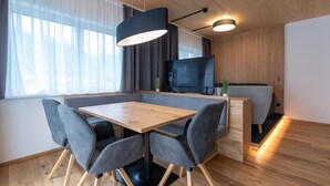A modern dining area with a wooden table and grey upholstered chairs, a black pendant light, and a television on a wooden wall. - A CASA Elegance (Laengenfeld)