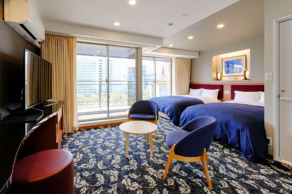 Ein Hotelzimmer mit zwei Betten, einem Fernseher, einem Schreibtisch, einem Stuhl und einem großen Fenster mit Blick auf die Stadt.