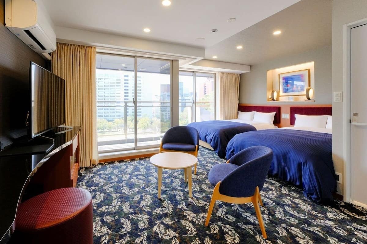 A hotel room with two beds, a TV, a desk, a chair, and a large window with a city view.