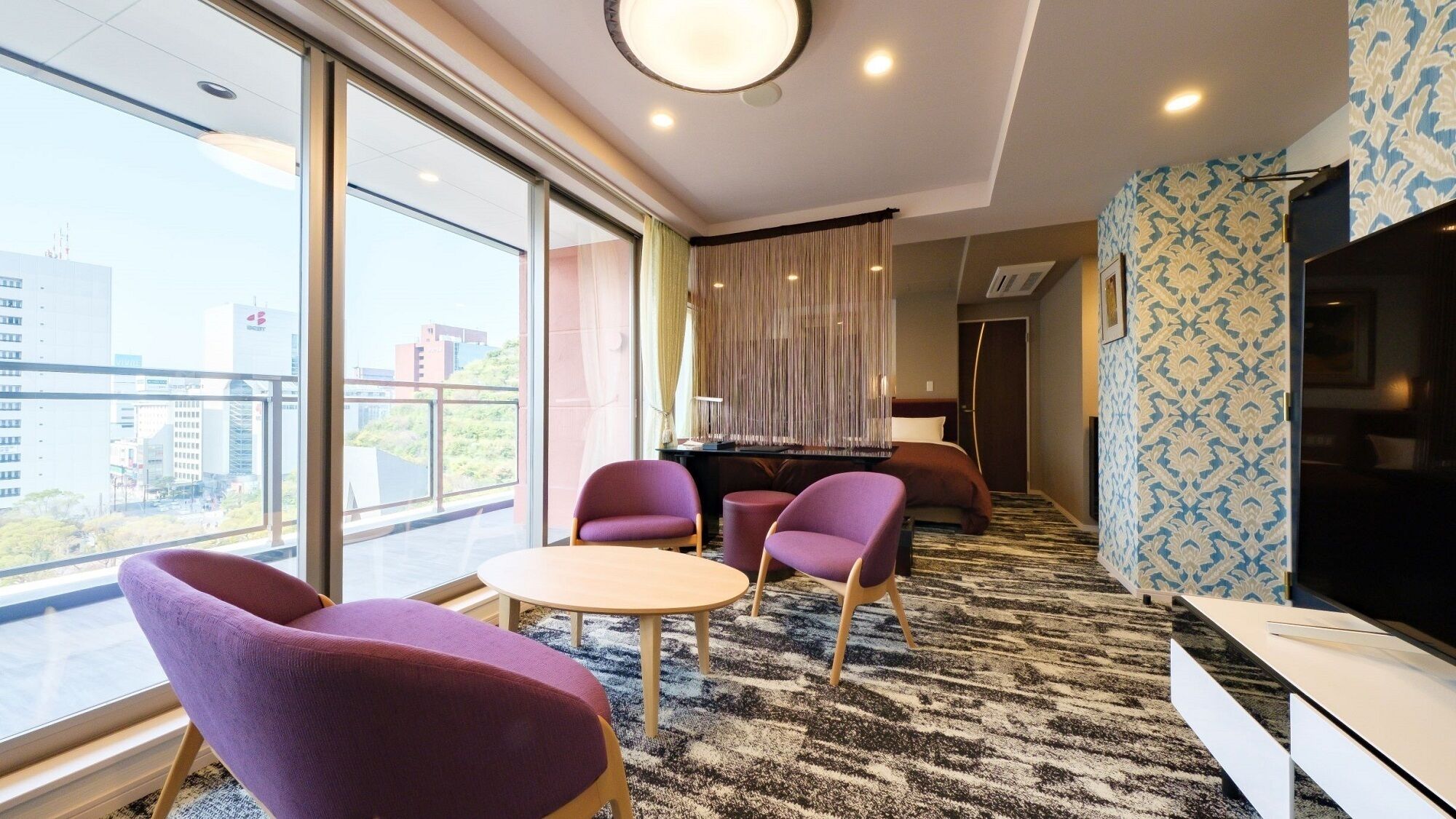 A modern hotel room with a large window, a round table, purple chairs, a TV, and a patterned wall.