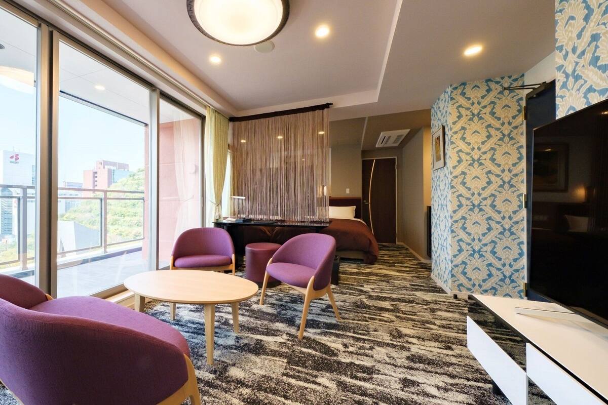 Ein modernes Hotelzimmer mit großem Fenster, einem runden Tisch, violetten Stühlen, einem Fernseher und einer Wand mit Muster.