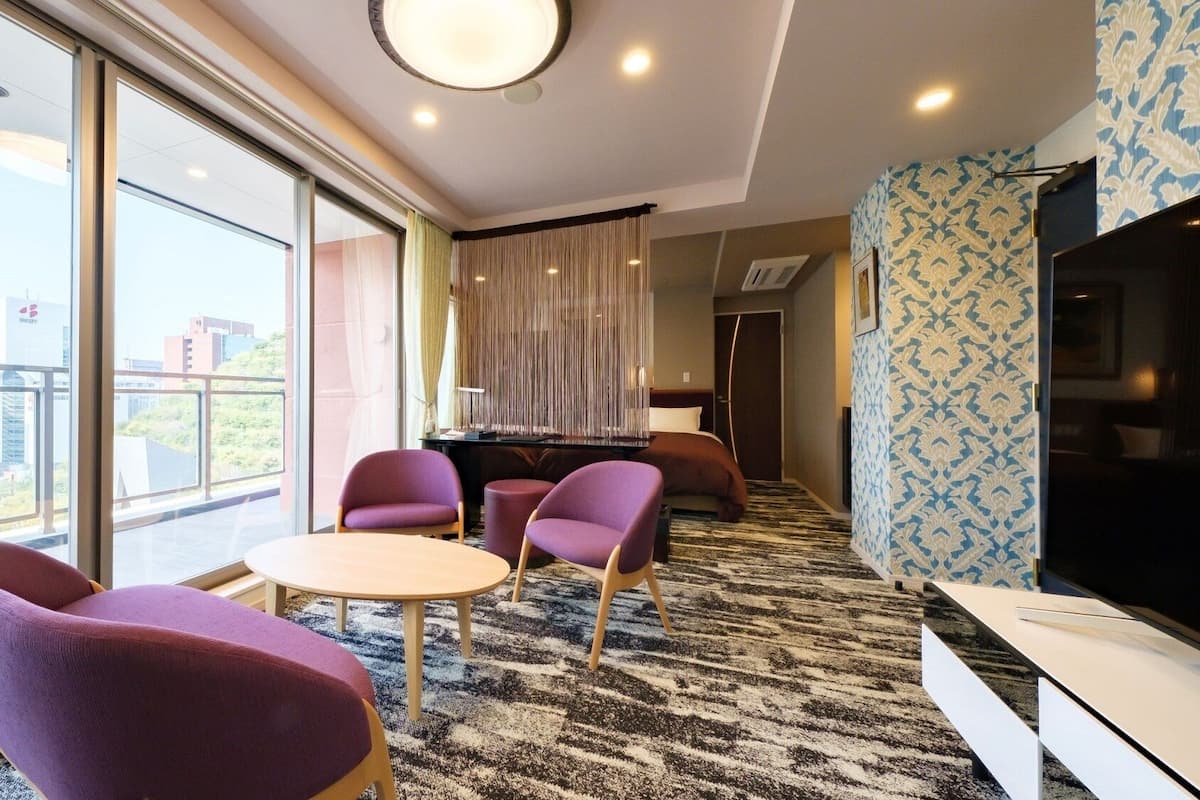 A modern hotel room with a large window, a round table, purple chairs, a TV, and a patterned wall.